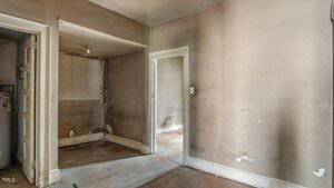 An empty, run-down interior room with stained and discolored walls, scuffed floors, visible damage, and a doorway leading to another room. A light bulb hangs from the ceiling in an adjacent alcove.