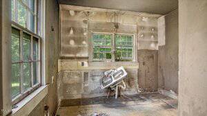 A small, neglected room with stained walls, a damaged floor, and an old sink tilted at an angle beneath two windows. The room appears rundown, with visible dirt, wear, and missing fixtures.