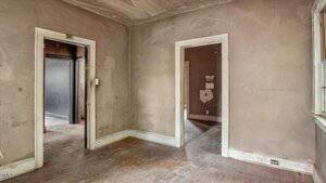 An empty, worn room with stained beige walls, scuffed wooden floors, two doorways leading to other rooms, and a window on the right letting in natural light. The trim and ceiling show signs of age and discoloration.