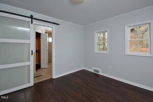 A small empty room with light gray walls, dark wood flooring, two windows, and a sliding frosted glass door partially open to a tiled area with a white door.