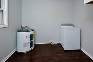 A small laundry room with a water heater on the left, a white washing machine on the right, wood-style flooring, light gray walls, and a window on the far left.