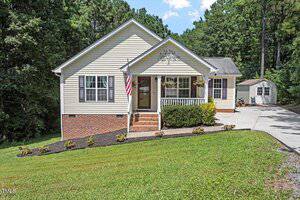 A single-story beige house with white trim at 4080 Brooksdale Drive, Franklinton. Features a front porch with an American flag, set on a sloped lawn with a driveway leading to a shed, all surrounded by trees.