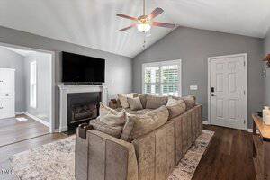 A living room with a sectional sofa, a wall-mounted TV above a fireplace, wood flooring, a ceiling fan, a rug, and a window with white shutters next to a white front door.