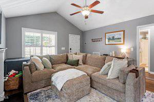 A living room with gray walls, a beige sectional sofa with multiple pillows, a ceiling fan, a window with shutters, a framed painting, and an open doorway leading to another room. A basket with toys is beside the fireplace.