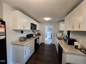A galley-style kitchen with white cabinets, dark countertops, and dark wood floors. Appliances including a microwave, toaster, and coffee maker are on the counters. A laundry room is visible at the far end.