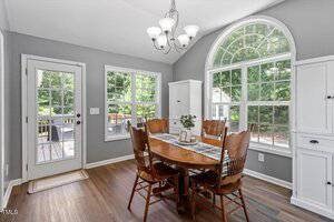 A dining area with a wooden table and five chairs, large windows, gray walls, white cabinetry, wood flooring, and a glass door leading outside. A chandelier hangs above the table. Trees are visible through the windows.