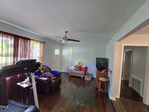A living room with dark wood floors, a treadmill, a recliner with a blue blanket, a loveseat with plush toys, a ceiling fan, a TV on a small table, and a window with sheer red curtains.