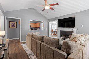 A living room with gray walls, a ceiling fan, a large beige sectional sofa, a flat-screen TV mounted above a fireplace, and a view into a kitchen and dining area through open doorways.