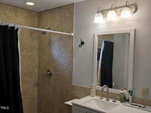 A bathroom with beige tiled shower, silver fixtures, white vanity with sink, mirror, soap, and toiletries. Wall-mounted lights above mirror, black shower curtain, and a single hook on the wall.