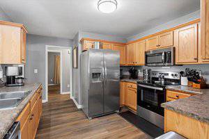 A modern kitchen with wooden cabinets, stainless steel appliances including a refrigerator and oven, dark countertops, a double sink, and wood-look flooring. There is a doorway leading to another room in the background.