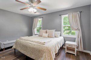 A bedroom with gray walls, a ceiling fan, two windows with light curtains, a bed with neutral bedding and throw pillows, a white nightstand, and a small bench next to the bed on wood flooring.