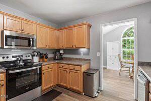 A kitchen with light wood cabinets, stainless steel appliances, cookware on the counter, a coffee maker, and a trash can. An adjacent dining area with large windows and a table is visible through the doorway.