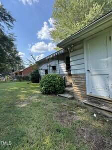 A weathered house with white siding and stone accents, featuring a bush near the entrance, sits on a grassy yard under a partly cloudy sky. The door and exterior appear worn and in need of maintenance.