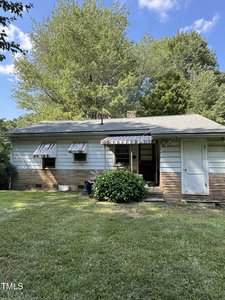 A small, single-story house with light blue siding and stone accents, two metal awnings over side windows, an awning over the back door, and a grassy yard surrounded by trees under a blue sky.