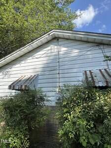 The exterior of a white house with horizontal siding, two windows with striped awnings, overgrown bushes in front, and a power line running vertically up the side wall. The sky is partly cloudy.