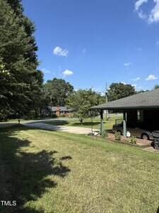 A grassy yard with a paved driveway leading to a house partially visible on the right; a car is parked under a carport. Another house and trees are visible in the background under a clear blue sky.