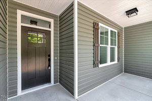 A green vinyl-sided house exterior with a black front door, a rectangular transom window above the door, white trim, a window with brown shutters, and a covered concrete porch.