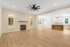 Spacious open-concept living area with light wood flooring, a ceiling fan, fireplace, and large windows. The area connects to a modern kitchen with an island, pendant lights, and white cabinetry.