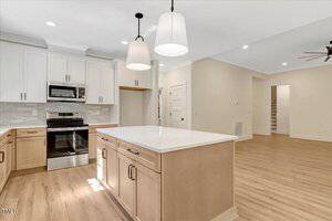Modern kitchen with light wood floors, white cabinets, stainless steel appliances, an island with a white countertop, and pendant lights. The space opens to a hallway and stairs with neutral-colored walls.