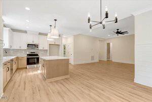 A spacious modern kitchen with light wood floors, white cabinets, an island with pendant lights, stainless steel appliances, and an open living area with a chandelier and ceiling fan. Stairs are visible in the background.