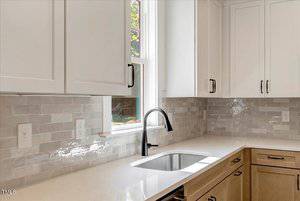 A modern kitchen features light wood lower cabinets, white upper cabinets, a stainless steel sink with a black faucet, white quartz countertops, and glossy light gray subway tile backsplash beneath a window.