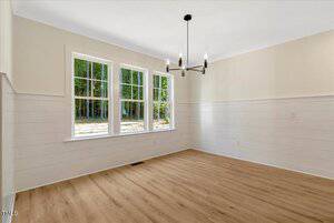 A bright, empty room with light wood floors, white shiplap walls, three large windows, and a modern black chandelier hanging from the ceiling. Sunlight streams through the windows, revealing a view of trees outside.
