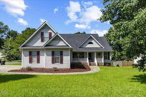 A gray single-family house with dark shutters, a covered front porch, and a gabled roof sits on a well-maintained green lawn with mulch beds and young shrubs. Trees and a wooden fence are visible in the background.