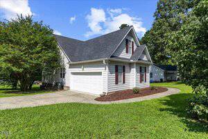 A white, two-story house with a dark gray roof and attached two-car garage sits on a green lawn. A curved driveway leads to the garage. Trees and shrubs line the property, and the sky is partly cloudy.