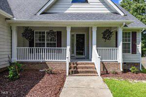 A front porch with white railings and columns, brown brick steps, two windows with shutters, and hanging plants. The yard is mulched with small shrubs and a concrete walkway leading to the steps.