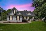 A gray two-story house with dark shutters and a covered front porch sits on a well-maintained lawn at 2735 Clifton Avenue in Creedmoor. A curved walkway leads to the entrance. The pink and blue sunset sky frames trees in the background.