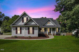 A gray two-story house with dark shutters and a covered front porch sits on a well-maintained lawn at 2735 Clifton Avenue in Creedmoor. A curved walkway leads to the entrance. The pink and blue sunset sky frames trees in the background.