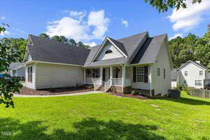 Single-story white house with dark roof, front porch with columns, hanging plants, and a grassy yard, set against a backdrop of trees and neighboring homes under a partly cloudy sky.