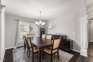 A dining room with a dark wood table and six beige upholstered chairs on a patterned rug, a chandelier overhead, a large window with curtains, and a dark sideboard and lamp against a light gray wall.