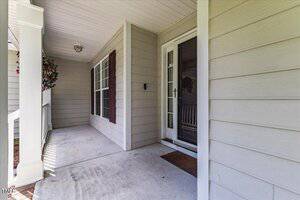 A covered front porch with light-colored siding, large windows with dark shutters, a brown doormat, and a glass storm door. A hanging planter is visible near the window, and the porch has a white railing.