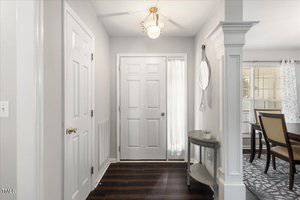 A small entryway with a white front door, a round ceiling light, dark wood flooring, a narrow table against the wall, and a dining area visible to the right. Light streams through sheer curtains.