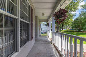 A covered front porch with white railing, columns, hanging plant, and large windows overlooks a green lawn and suburban street with trees and houses visible in the background.