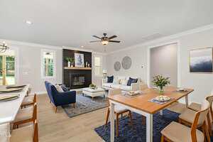 A modern open-concept living and dining area with a fireplace, blue and white furniture, wooden dining table with chairs, and decorative accents on the walls and table. Light wood flooring and natural light from windows.