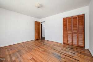 An empty room with white walls, a wooden floor, an open wooden door, and a closed closet with louvered wooden doors. A ceiling light fixture is centered above.