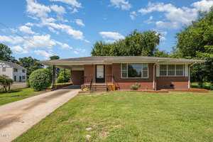 A single-story brick house with a carport on the left, a concrete driveway, front steps, a white door, large windows with white bars, and a grassy front yard under a blue sky with scattered clouds.