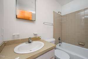 A bathroom with a beige countertop, white sink, wall-mounted mirror, toilet, and a bathtub with beige tile surround. Two empty soap holders are above the sink, and a towel rack is on the wall next to the tub.