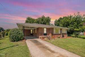 A single-story brick house at 309 W Rockspring St. in Henderson features a covered carport, front porch, and driveway, surrounded by grass, bushes, and trees under a colorful sunset sky.