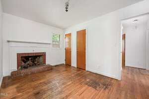 A small, empty room with white walls, a brick fireplace, hardwood floors, two wooden doors, a window, and a hallway entrance. The floor shows signs of wear and there is a light fixture on the ceiling.