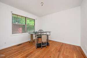 A mostly empty room with hardwood floors, white walls, a single window with security bars, and a corner desk with a chair. The ceiling has an exposed light fixture without a cover.