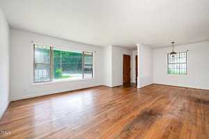 Unfurnished living and dining area with white walls, wood floors, two large windows, a hanging light fixture, and a wooden front door. The space is open and bright with natural light.