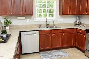 A kitchen with brown wooden cabinets, a stainless steel dishwasher, a double sink under a window, beige countertops, and a gray rug on a tiled floor. A stand mixer and potted plant are on the counter.