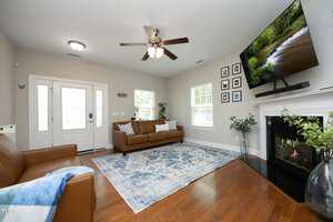 A living room with brown leather sofas, a blue patterned rug, hardwood floors, a ceiling fan, a fireplace with a TV mounted above it, plants, and framed pictures on the wall near a white-framed window.