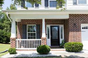 A brick house with a small front porch featuring white columns, a black front door with glass panels, two windows with dark shutters, and neatly trimmed shrubs in front.