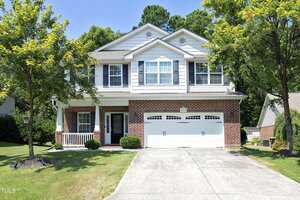 Charming two-story suburban house at 2019 Massimo Drive in Creedmoor, featuring red brick and white siding, double garage doors, a concrete driveway, well-kept lawn, small shrubs near the entrance, and trees at the front and sides.