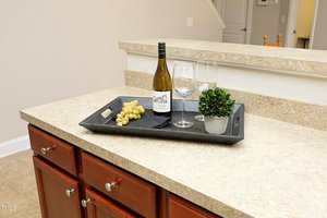 A kitchen counter with a black tray holding a bottle of white wine, two empty wine glasses, a small bunch of green grapes, and a small potted plant. The counter has a beige surface and wooden cabinets underneath.
