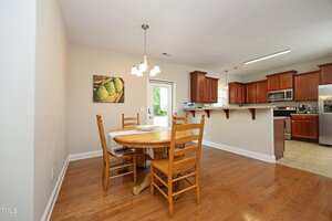 A dining area with a round wooden table and four chairs on a wood floor, next to a kitchen with stainless steel appliances and dark wood cabinets. A pendant light hangs above the table, and a painting is on the wall.